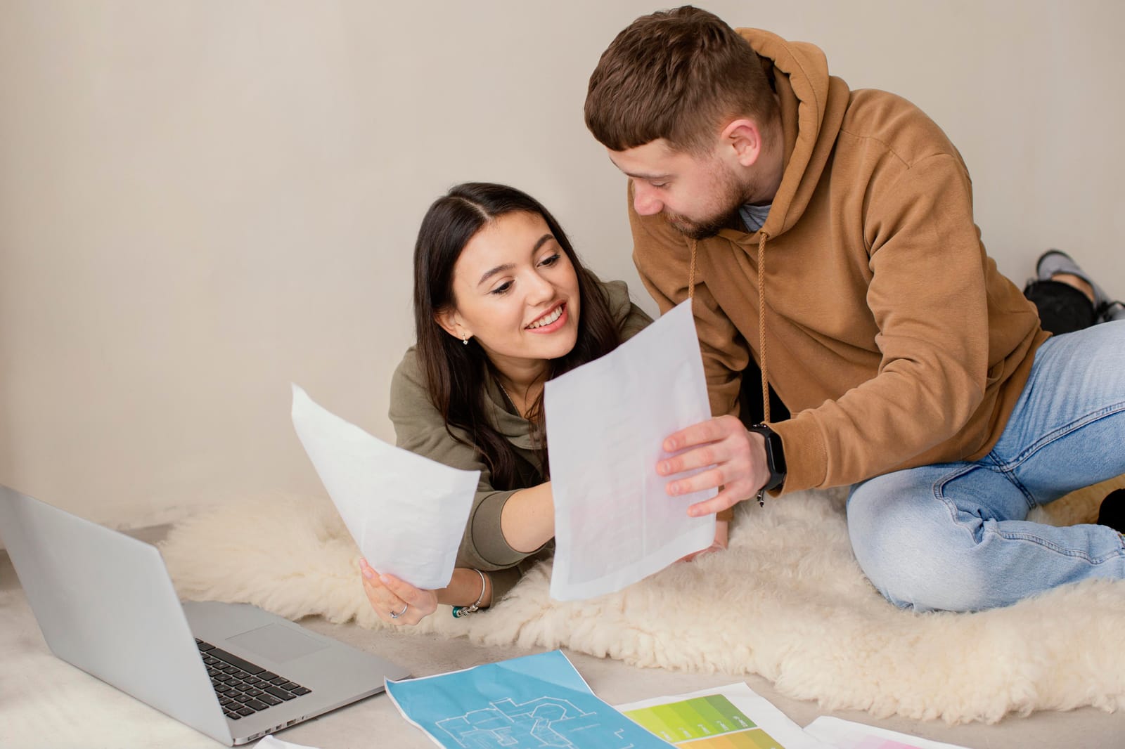 A young couple reviews housing documents and floor plans beside a laptop while discussing their next apartment search.