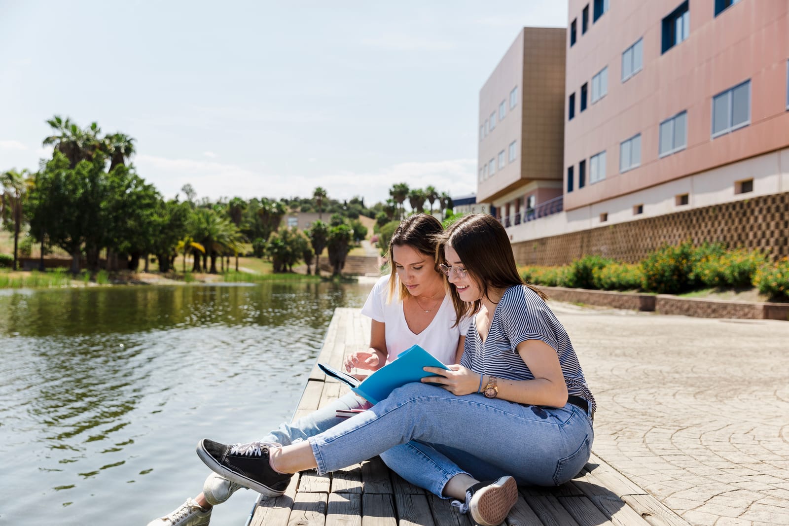 Two college students sit by a campus pond, reviewing notes together outdoors near a university building on a sunny day.